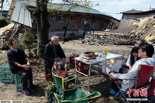 当地时间4月17日，日本熊本县震后见闻。一家人围坐在简易桌旁吃饭，周围都是倒塌的房屋。