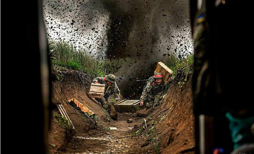 遭到炮击的乌军堑壕，两名士兵正冒着炮火搬运弹药箱。